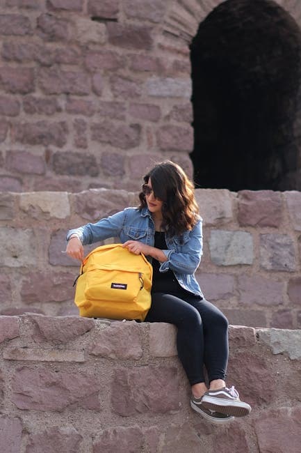 A woman sits on a historic stone wall in Ankara, checking her backpack on a sunny day.